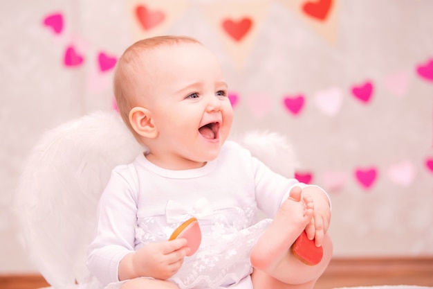 Portrait of a cheerful little girl with white feather wings eating heart-shaped cookies