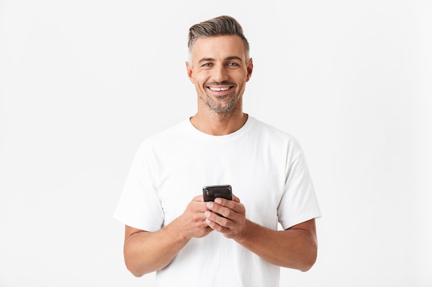 Portrait of cheerful man 30s wearing casual t-shirt holding and using smartphone isolated on white