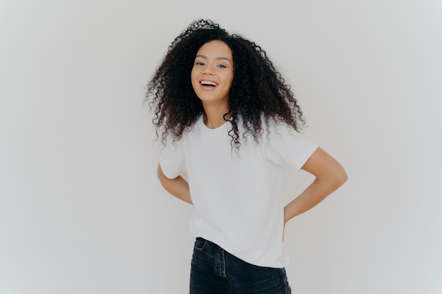 Portrait of cheerful millennial woman keeps hands behind back dressed in casual wear laughs from something fun isolated over white studio background African American lady expresses happiness