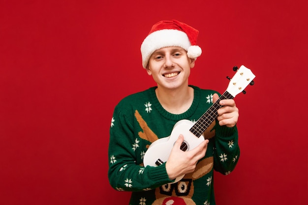 Portrait cheerful musician in santa hat on red background playing on ukulele