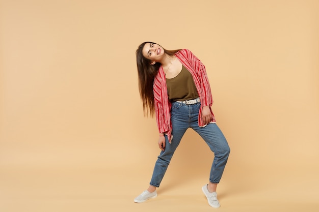 Portrait of cheerful pretty young woman in casual clothes standing, looking camera isolated on pastel beige wall background in studio. People sincere emotions, lifestyle concept. Mock up copy space.