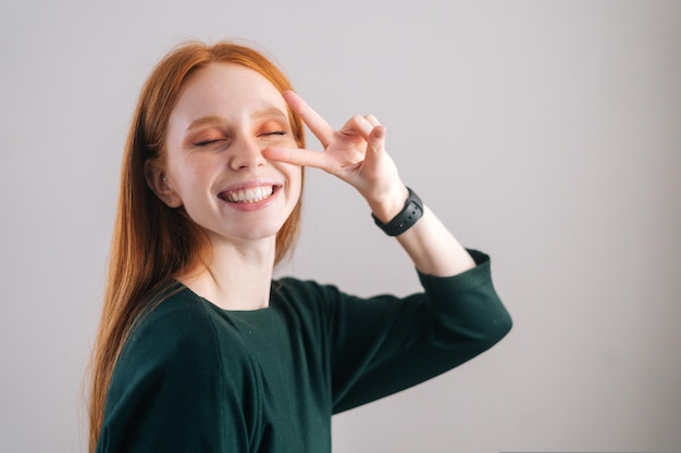 Portrait of cheerful redhead young woman model showing victory sign with fingers eyes closed on