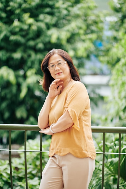 Portrait of cheerful senior Vietnamese woman in glasses standing outdoors on balcony and smiling at camera