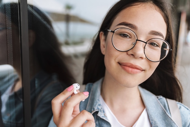 Portrait of a cheerful smiling young pretty woman student wearing eyeglasses walking outdoors resting listening music with earphones