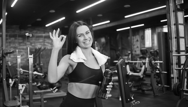 Portrait of a cheerful smilling young sportswomanin in sportswear and towel around his neck, bottle of water in hand shows fingers ok sign in gym. Positive emotions, training process