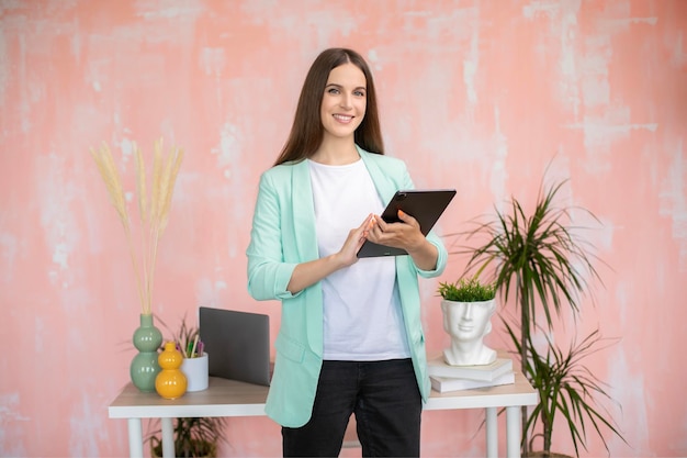 Portrait of cheerful stylish girl in smart casual outfit standing at desk holding tablet in hands