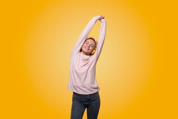 Portrait of a cheerful stylish young woman standing isolated over yellow background listening to music with headphones dancing