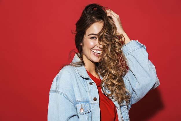 Portrait of a cheerful stylish young woman wearing denim jacket standing isolated over red wall, posing