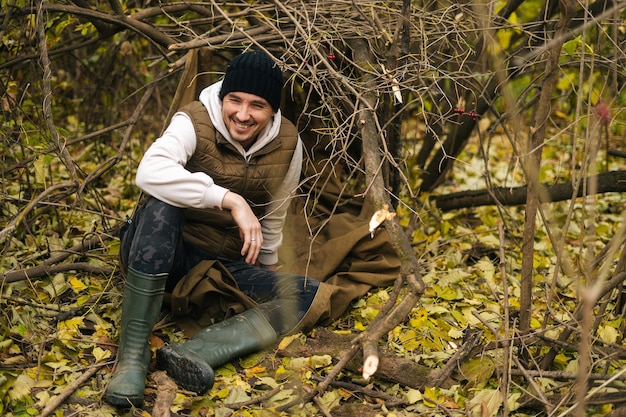 Portrait of cheerful tourist man sitting on spread out raincoat in thicket under tree in woods on overcast rainy day to rest