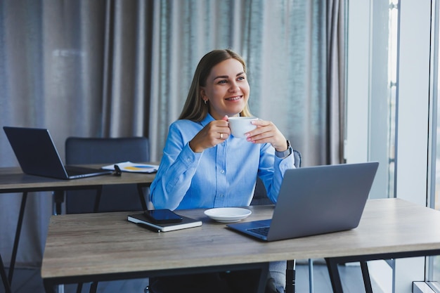 Portrait of cheerful woman in classic glasses smiling in free time in cafe with coffee positive european woman in blue shirt desk with laptop remote work