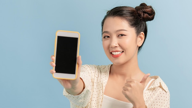 Portrait of a cheerful woman showing blank smartphone screen and showing thumb up isolated on a turquoise background