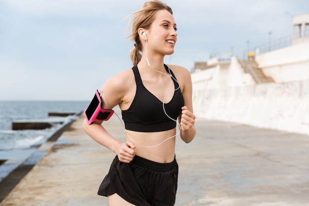 Portrait of cheerful woman wearing smartphone case listening to music with earphones while jog near seaside in morning