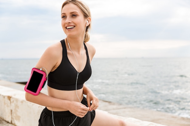 Portrait of cheerful woman wearing smartphone case listening to music with earphones while working out near seaside in morning