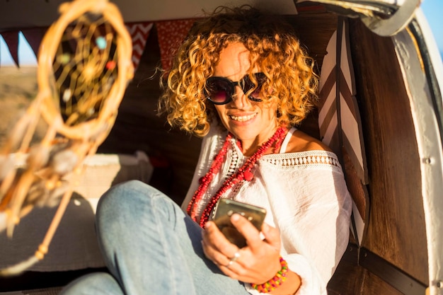 Portrait of cheerful young adult woman using phone sitting and enjoying inside classic travel van vehicle Pretty female people with connection cellular technology and planning road trip for vacation