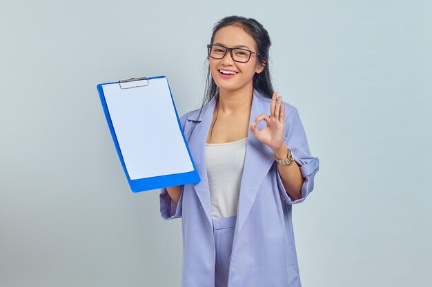 Photo portrait of cheerful young asian business woman holding blank clipboard and showing ok gesture isolated on purple background