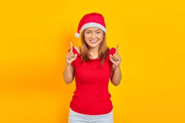 Portrait of cheerful young Asian woman pointing up while wearing Christmas hat on yellow background