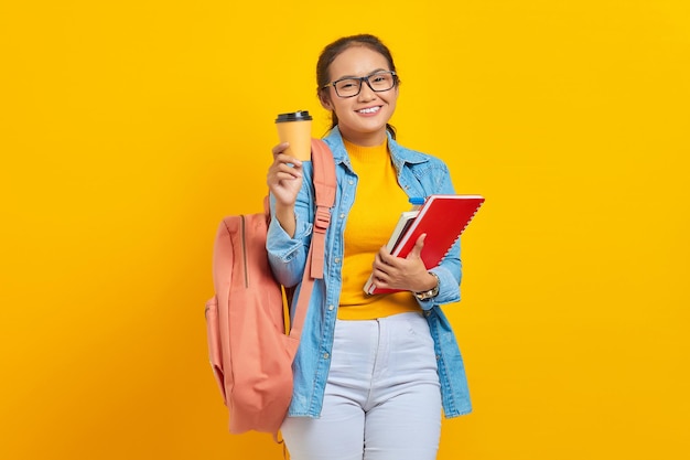 Portrait of cheerful young Asian woman student in casual clothes with backpack holding book and showing coffee take away looking at camera isolated on yellow background