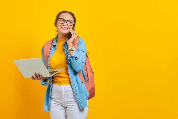 Portrait of cheerful young Asian woman student in casual clothes with backpack talking on mobile phone and holding laptop isolated on yellow background. Education in college university concept