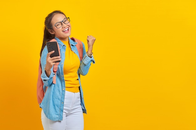 Portrait of  cheerful young Asian woman student in casual clothes with backpack using mobile phone and celebrating success, getting good news isolated on yellow background