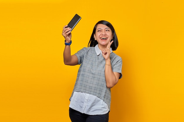 Portrait of cheerful young Asian woman using mobile phone take selfie make peace sign