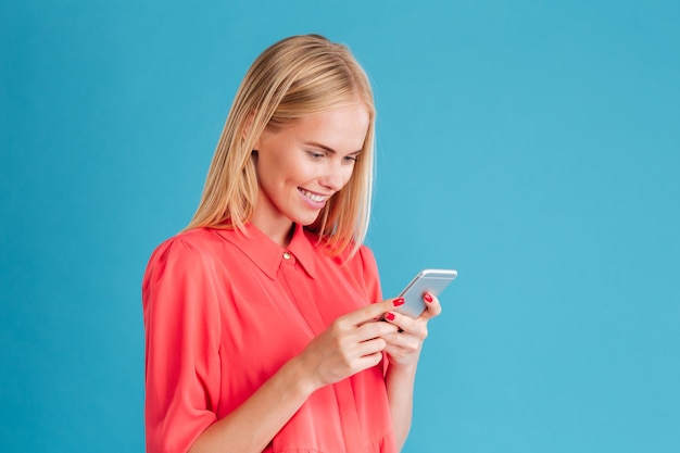 Portrait of a cheerful young blonde woman holding mobile phone over blue wall