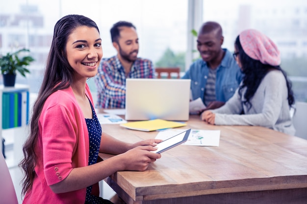 Portrait of cheerful young businesswoman holding digital tablet while sitting with colleagues in office