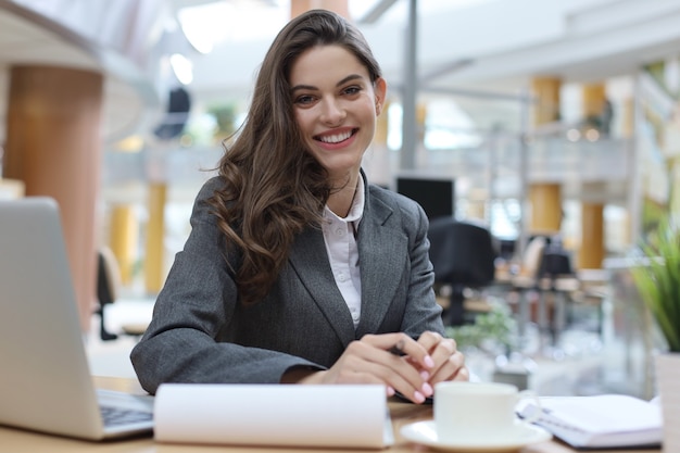 Portrait of a cheerful young businesswoman sitting at the table in office and looking at camera.