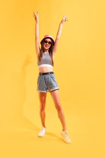 Portrait of cheerful young girl in pink panama sunglasses and summer outfit posing isolated over yellow studio background