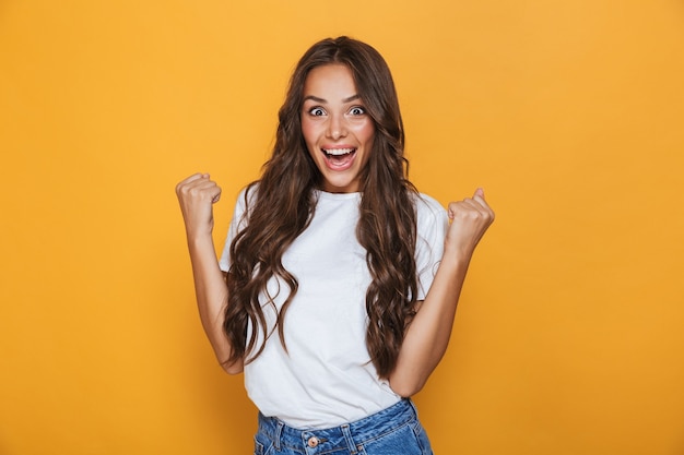 Portrait of a cheerful young girl with long brunette hair standing over yellow wall, celebrating success