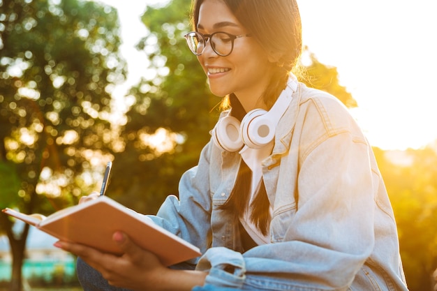 Portrait of a cheerful young student girl wearing eyeglasses sitting outdoors in nature park listening to music with headphones and writing notes