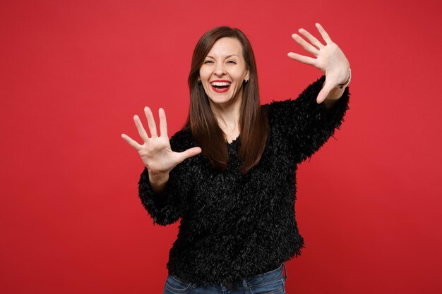 Portrait of cheerful young woman in black fur sweater standing showing palms on camera isolated on bright red wall background in studio. People sincere emotions, lifestyle concept. Mock up copy space.