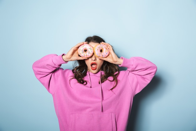 Photo portrait of cheerful young woman with two ponytails smiling while holding sweet donuts over blue in studio