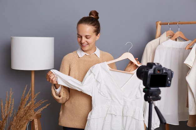 Photo portrait of cheery young girl recording her video blog episode about new white dress shoes while being at home female in beige jumper with bun hairstyle