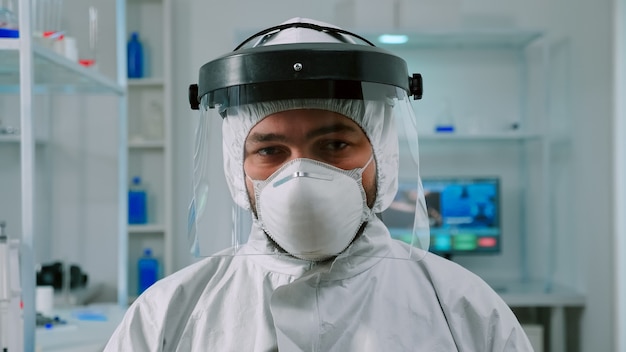 Portrait of chemist doctor looking up at video camera sitting in modern equipped lab wearing protection suit. Examining virus evolution using high tech and chemistry tools for vaccine development.