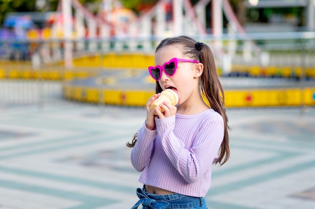 Portrait of a child girl in an amusement park in the summer eating ice cream near the carousels and smiling with happiness in sunglasses the concept of weekends and school holidays