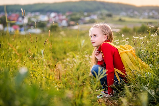 Portrait of a child girl in a red jumper with a backpack traveling on a summer evening against the backdrop of a landscape.