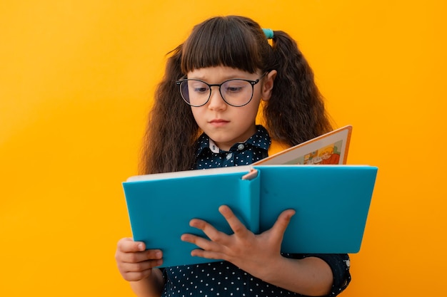 Portrait of a child schoolgirl with glasses reading a book on an isolated yellow background