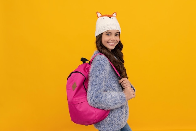 Portrait of child wearing warm clothes carry backpack. express positive emotion. winter fashion. smiling kid with curly hair in hat. back to school. teen girl in knitwear on yellow background.