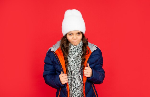 Photo portrait of child wearing warm clothes with scarf. express emotion. winter fashion. naughty kid with curly hair in puffer jacket and hat. teen girl on red background.