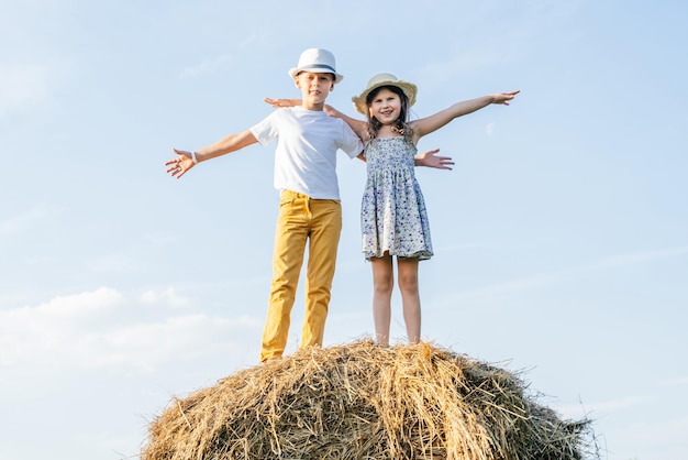 Portrait of children boy and girl spreading and waving arms smiling on haystack in field Wearing straw hats