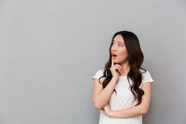 Portrait of chinese surprised woman with dark curly hair posing on camera and looking aside on copyspace with finger on lips, isolated over gray wall