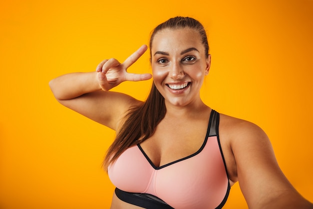 Portrait of chubby sportswoman in sportive bra using mobile phone for selfie photo, isolated over yellow background