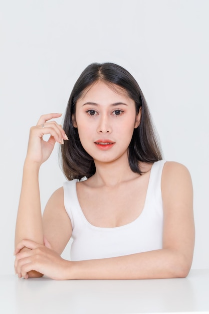 Portrait close up studio shot of millennial Asian short black hair model wearing makeup red lip in tank top undershirt sitting at table posing holding hand on cheek look at camera on white background.