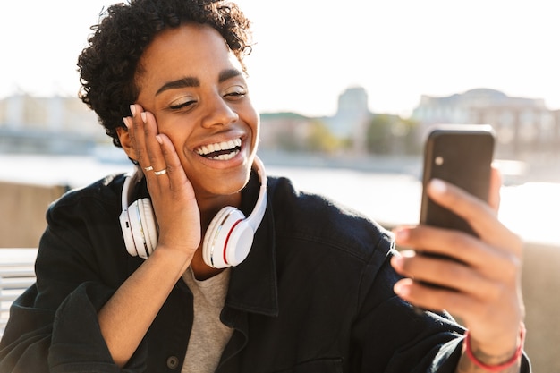 Portrait closeup of friendly woman with headphones making video call on smartphone while sitting on bench near riverfront