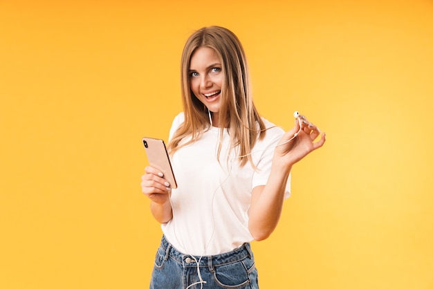 Portrait closeup of positive blond woman smiling while listening to music with smartphone and earphones isolated over yellow wall