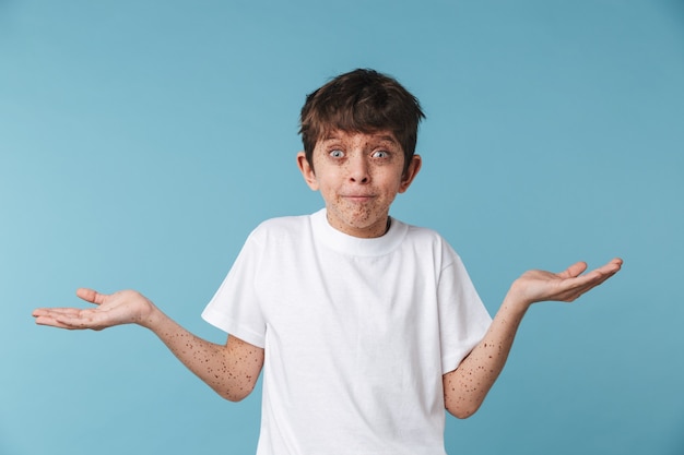 Portrait closeup of puzzled beautiful boy with freckles wearing white casual t-shirt looking at front isolated over blue wall