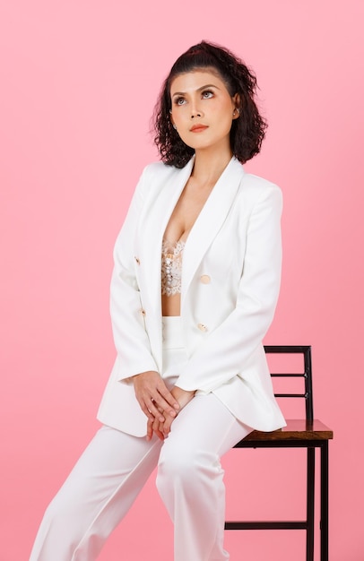 Portrait closeup studio shot of Asian sexy curly hairstyle successful businesswoman in white fashionable casual suit with lace lingerie crop top bra sitting on tall chair posing on pink background