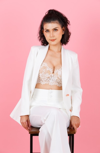 Portrait closeup studio shot of Asian sexy curly hairstyle successful businesswoman in white fashionable casual suit with lace lingerie crop top bra sitting on tall chair posing on pink background