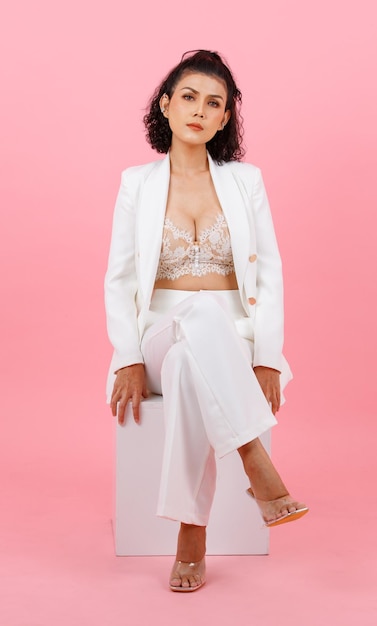 Portrait closeup studio shot Asian young sexy curly hairstyle professional successful businesswoman in white fashion casual suit and lace bra sitting smiling crossed legs posing on pink background