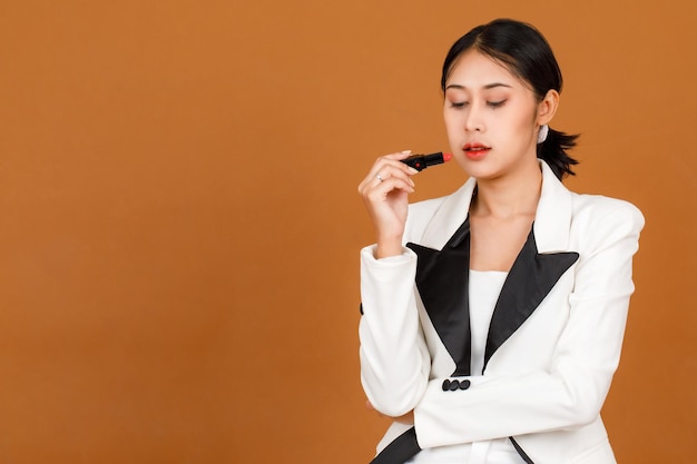 Portrait closeup studio shot millennial Asian black short ponytail hair female model wears makeup in black and white fashion casual blazer crossed arm holding red lipstick in hand on brown background.